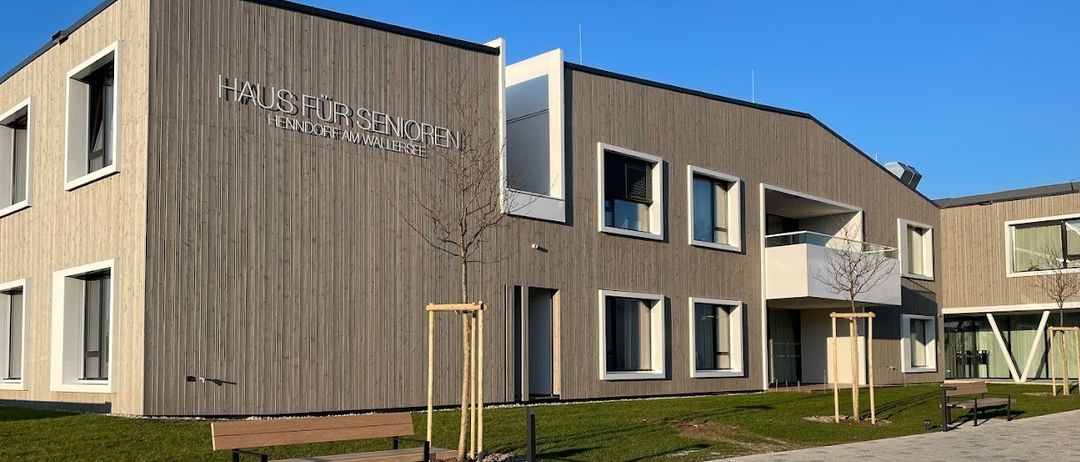 Ein modernes Gebäude mit dem Schild 'Haus für Senioren' davor, mit einer Holzbank und einem kleinen Baum davor.