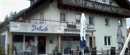 Ein weißes Gebäude mit Balkon und Schild mit der Aufschrift DorCafe, mit Sitzgelegenheiten im Freien und Tischen unter Schirmen.