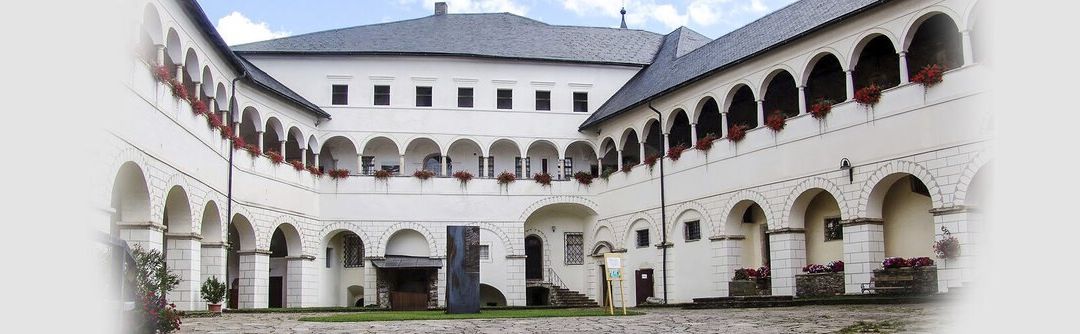 Der Innenhof eines historischen Schlosses mit Bogenfenstern und roten Blumen in hängenden Töpfen. Der Hof ist von Steinmauern umgeben und hat einen gepflasterten Boden.