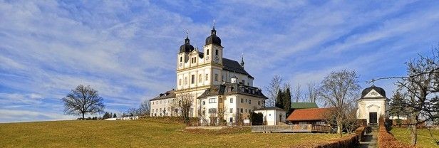 Eine große Kirche mit zwei Kuppeln steht auf einem grasbewachsenen Hügel unter einem teilweise bewölkten Himmel, umgeben von Bäumen und einer kleinen Kapelle.