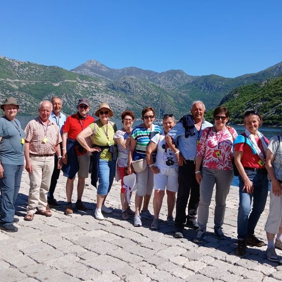Eine Gruppe von Touristen steht auf einem Steinweg und trägt Hüte und Sonnenbrillen. Sie lächeln für ein Foto mit Bergen im Hintergrund.