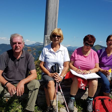 Vier Personen sitzen auf einer Steinbank in einer Bergregion. Zwei Frauen schreiben in Notizbüchern, die anderen beiden beobachten. Sie tragen alle Sonnenbrillen.