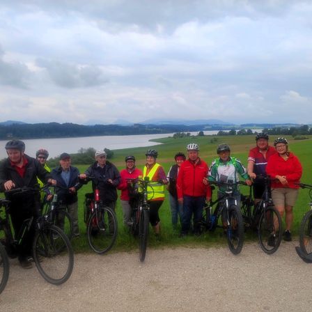 Eine Gruppe von Menschen posiert für ein Foto auf einem Weg mit ihren Fahrrädern, vor einem See und grünen Hügeln.