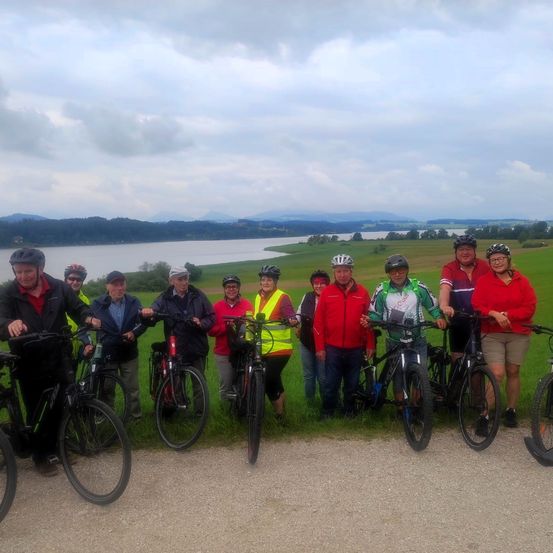 Eine Gruppe von Menschen posiert für ein Foto auf einem Weg mit ihren Fahrrädern, vor einem See und grünen Hügeln.