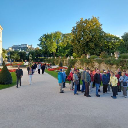 Eine Gruppe von Menschen steht in einem Garten mit blauem Himmel. In der Ferne befindet sich ein Schloss mit Turm und Bäumen.