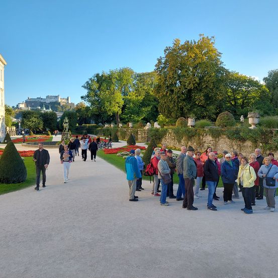 Eine Gruppe von Menschen steht in einem Garten mit blauem Himmel. In der Ferne befindet sich ein Schloss mit Turm und Bäumen.