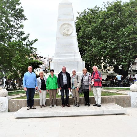 Eine Gruppe von Menschen steht vor einem Denkmal in einem Park mit Bäumen. Das Denkmal hat ein kreisförmiges Design mit einem Porträt. Die Menschen sind lässig gekleidet.