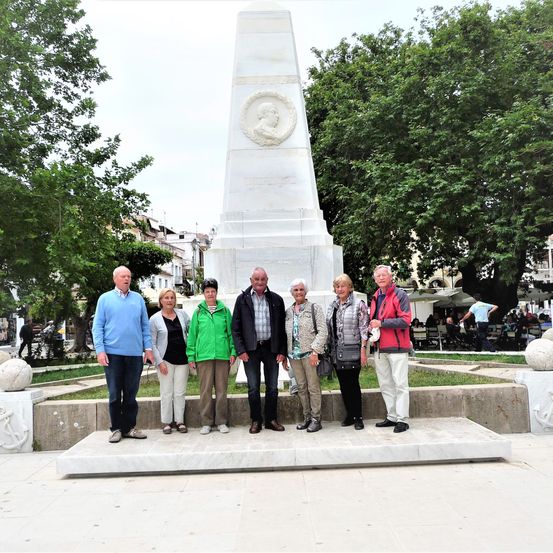 Eine Gruppe von Menschen steht vor einem Denkmal in einem Park mit Bäumen. Das Denkmal hat ein kreisförmiges Design mit einem Porträt. Die Menschen sind lässig gekleidet.