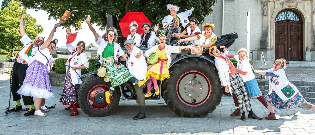 Eine Gruppe von Menschen in Clownkostümen posiert auf einem großen Traktor vor einem Gebäude mit einem Baum.