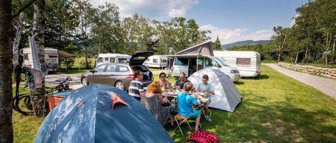Eine Familie sitzt um einen Tisch auf einer Wiese, wahrscheinlich auf einem Campingplatz.