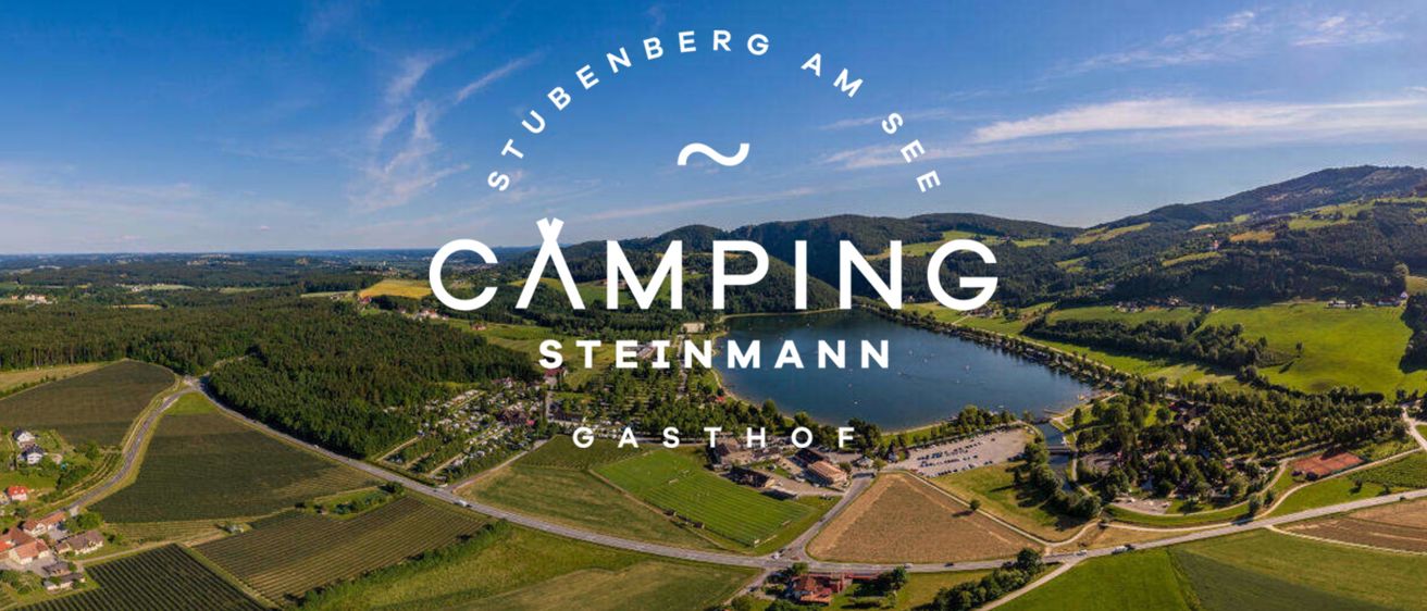Luftaufnahme des Camping Steinmann Gasthof, einer friedlichen Campinganlage, umgeben von üppiger Grünfläche und einem malerischen See, unter einem klaren blauen Himmel.