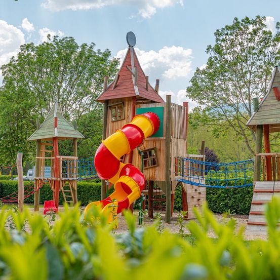 Ein Kinderspielplatz mit einer rot-gelben Spiralrutsche, Holztürmen und einer Seilleiter, vor dem Hintergrund von grünen Bäumen und Büschen unter einem bewölkten Himmel.
