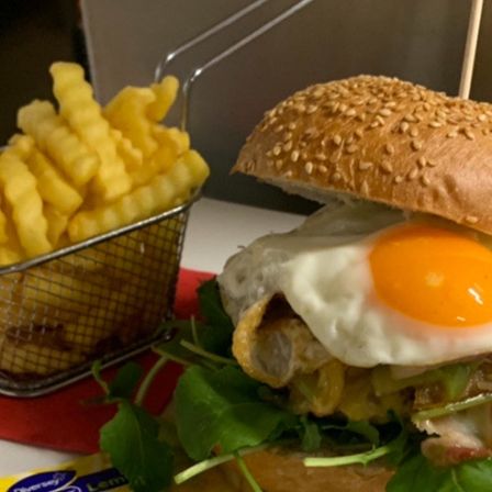 Ein Nahaufnahme eines Burgers mit einem Ei darauf neben einem Korb mit Pommes Frites.