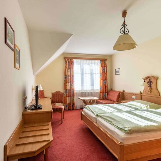 Ein Schlafzimmer mit einem Doppelbett, einem Schreibtisch und einem Stuhl. An der Wand hängen zwei gerahmte Bilder. Das Bett hat zwei Kissen und eine Decke. Eine Lampe hängt von der Decke.