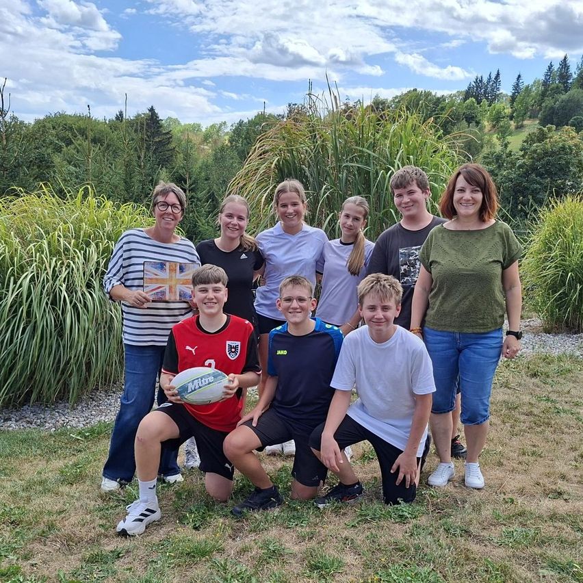 Eine Gruppe von Menschen, einschließlich Kindern und Erwachsenen, posiert für ein Foto in einem Grasbereich mit einem Rugbyball im Vordergrund.