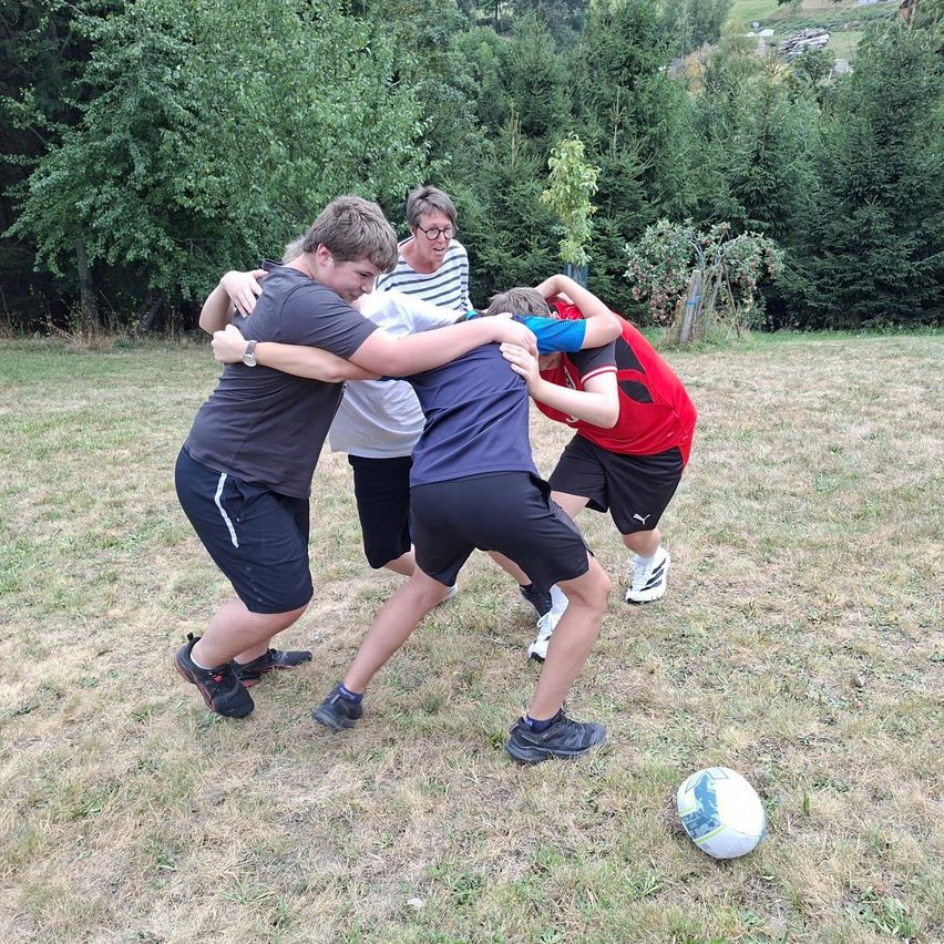 Vier Personen sind in einem Grasfeld in einen spielerischen Ringkampf verwickelt, mit einem Fußball in der Nähe.