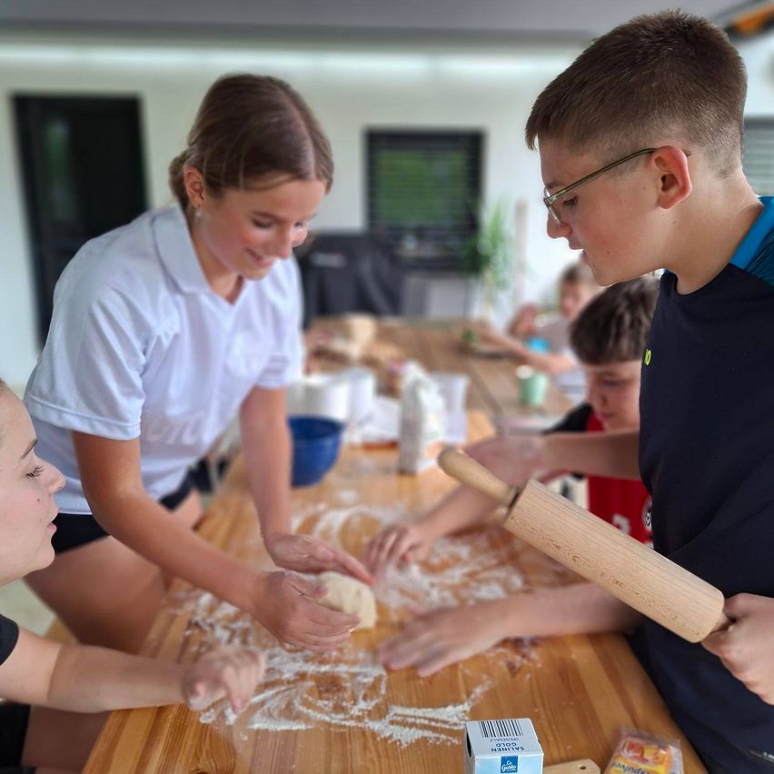 Eine Gruppe von Kindern und einer Frau macht Teig auf einem Holztisch. Die Frau lächelt und leitet die Kinder. Mehl ist auf dem Tisch verstreut. Ein Junge hält einen hölzernen Nudelholz.