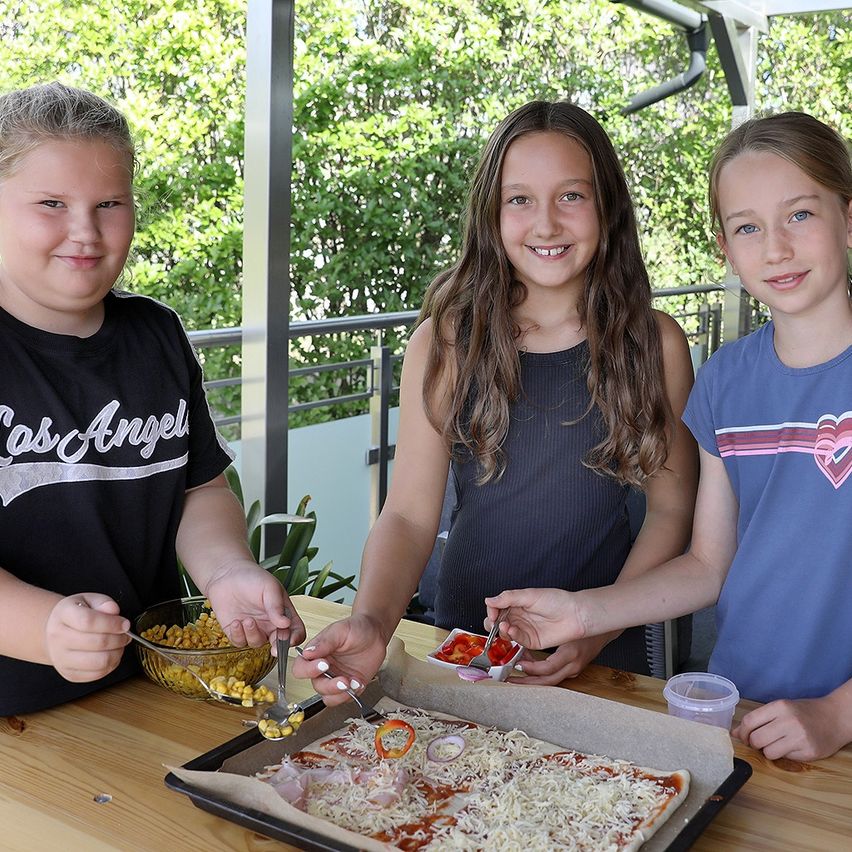 Drei junge Mädchen lächeln und fügen einer Pizza auf einem Holztisch Toppings hinzu. Eines der Mädchen fügt Mais hinzu, ein anderes Käse und das dritte Pepperoni.