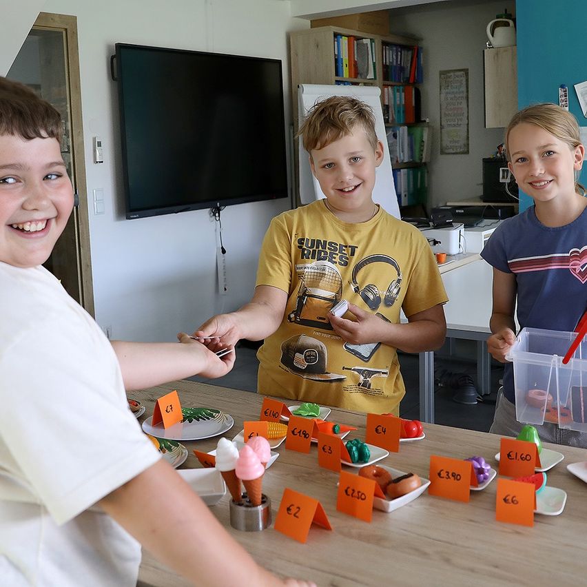 Drei Kinder stehen hinter einem Tisch mit verschiedenen ausgestellten Gegenständen. Der Junge auf der linken Seite lächelt und hält eine Karte. Der Junge in der Mitte hält eine Karte und schaut den Jungen auf der linken Seite an. Das Mädchen auf der rechten Seite hält einen Plastikbehälter.