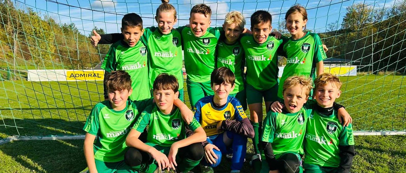 Eine Jugendfußballmannschaft posiert für ein Foto auf einem Feld. Sie tragen grüne und gelbe Trikots mit dem Wort 'mauk' darauf. Hinter ihnen steht ein Tornetz.