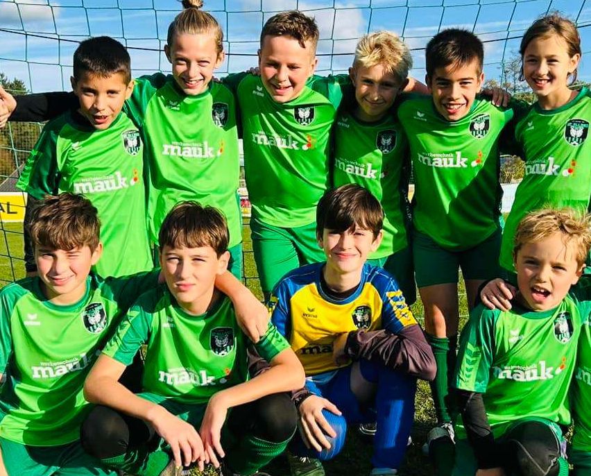 Eine Jugendfußballmannschaft posiert für ein Foto auf einem Feld. Sie tragen grüne und gelbe Trikots mit dem Wort 'mauk' darauf. Hinter ihnen steht ein Tornetz.