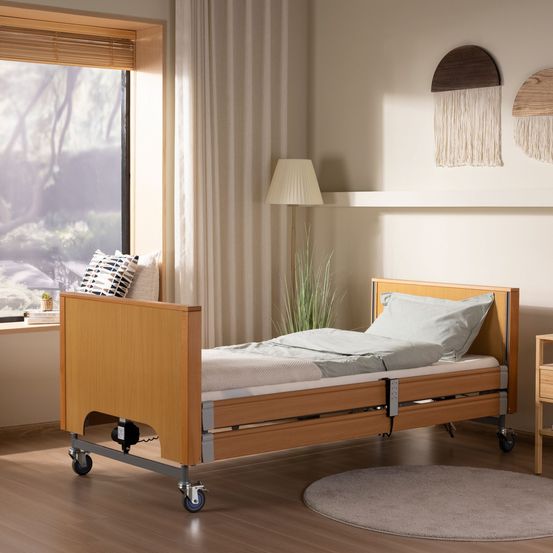 Ein Krankenhausbett mit Rädern steht in einem gut beleuchteten Raum. Es hat ein Kissen und eine Decke. In der Nähe gibt es eine Lampe und einen Teppich. Durch das Fenster ist eine Aussicht auf Bäume zu sehen.