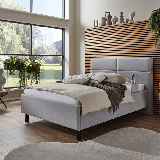 Ein modernes Schlafzimmer mit einem großen Fenster mit Blick auf eine Wiese. Ein graues Bett mit hölzerne Kopfstütze und Tagesdecke. Ein hölzerner Boden und eine Holzwand mit Dekorationen.