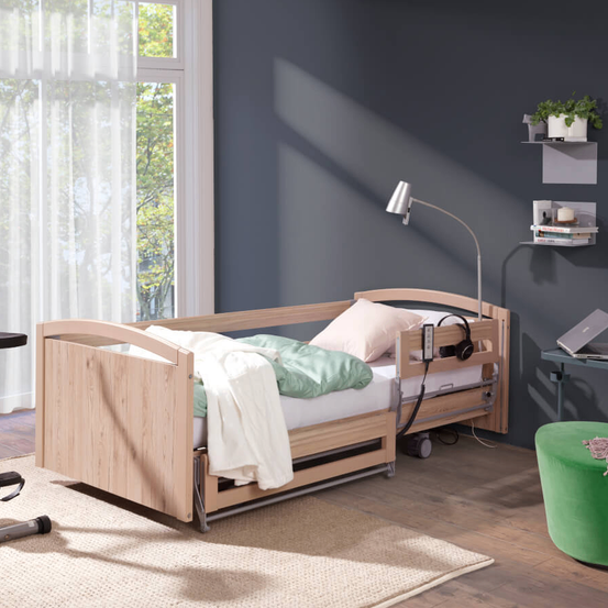 Ein Schlafzimmer mit einem Holzbett, Kissen und einer Nachttischlampe. Ein weißer Vorhang bedeckt das Fenster, draußen sind Bäume zu sehen.