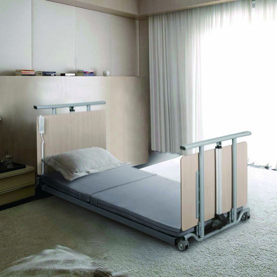 Ein kleines Krankenhauszimmer mit einem Bett, einem Nachttisch und einer Lampe. Der Raum hat einen Teppichboden, weiße Vorhänge und eine neutrale Farbpalette.