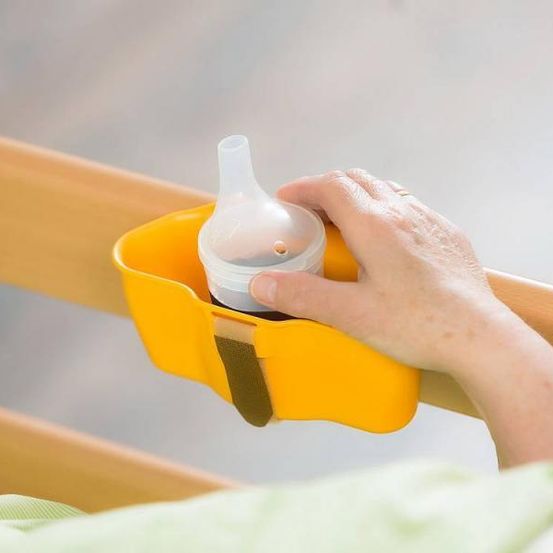 Eine Hand hält einen gelben Flaschenhalter an der Seitenschiene eines Krankenhausbettes. Die Flasche ist klar mit einem weißen Deckel. Der Arm der Person zeigt einige Sommersprossen.