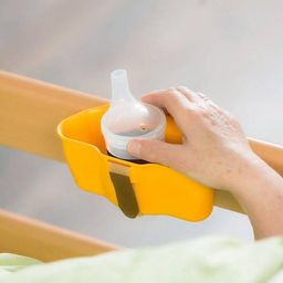 Eine Hand hält einen gelben Flaschenhalter an der Seitenschiene eines Krankenhausbettes. Die Flasche ist klar mit einem weißen Deckel. Der Arm der Person zeigt einige Sommersprossen.