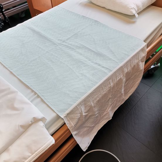 Ein Krankenhausbett mit einem blauen Matratzenschoner. Das Bett ist mit weißen Laken bedeckt und hat zwei weiße Kissen. Der Boden ist mit schwarzen Fliesen belegt.