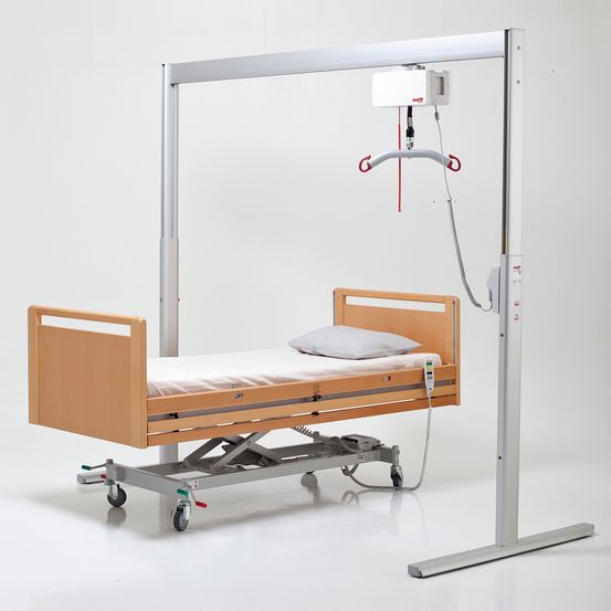 Ein elektrisches Krankenhausbett mit angeschlossenem Deckenliftsystem, einem Kissen und Rädern für Mobilität, vor weißem Hintergrund.