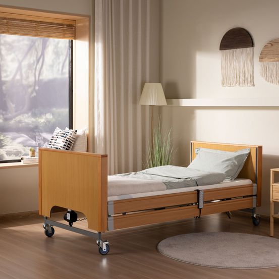 Ein Krankenhausbett mit Rädern steht in einem Raum mit Fenster und weisser Gardine. Ein weisses Kissen und eine Topfpflanze sind auf der Fensterbank. Eine Lampe und eine Pflanze befinden sich auf der rechten Seite.