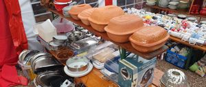 Eine Vielzahl von Küchenutensilien ist auf einem Holzregal ausgestellt, darunter Tontopf, Metallschalen und durchsichtige Plastikbehälter. Schüsseln, Teller und Tassen sind im Hintergrund gestapelt. Die Umgebung scheint ein Markt oder Verkauf mit einem Zelt im Hintergrund zu sein.