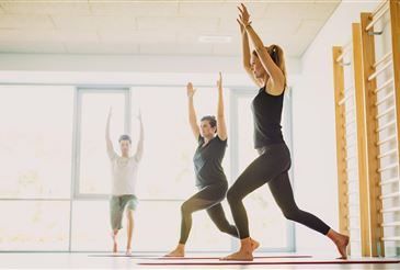 Drei Personen praktizieren Yoga in einem Raum mit einer Holzstange an der Wand, eine von ihnen vollführt eine Handstand.