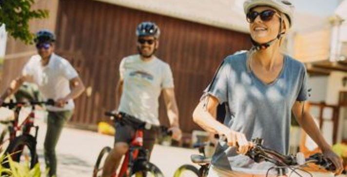 Drei Personen fahren auf einem Weg vor einer Scheune mit Fahrrädern. Alle tragen Helme und Sonnenbrillen. Die Frau vorne lächelt. Dahinter fahren zwei Männer, einer trägt ein weißes T-Shirt und der andere ein blaues.