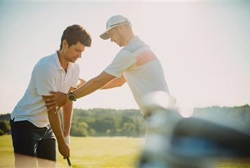 Zwei Männer sind auf einem Golfplatz; einer unterrichtet den anderen im Golfschwung. Der Ausbilder führt die Hände des Lernenden.