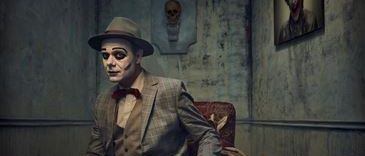 Ein Mann in einem Vintage-Anzug, einem Fedora und Gesichtsbemalung sitzt auf einem Stuhl in einem schwach beleuchteten Raum mit einem Schädel und einem Gemälde an der Wand.