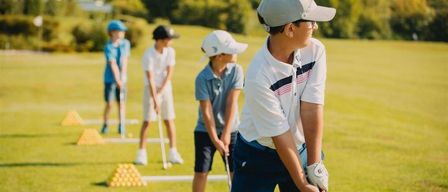 Eine Gruppe junger Jungen übt Golf an einem sonnigen Tag, mit einem Gebäude im Hintergrund und einem Hintergrund aus Bäumen.