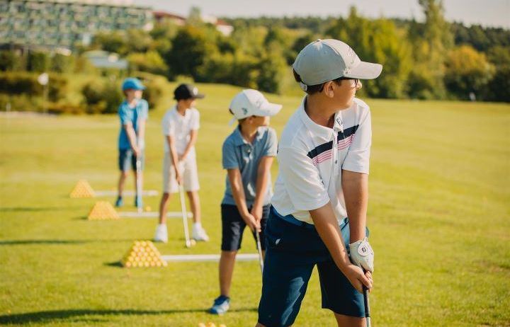 Eine Gruppe junger Jungen übt Golf an einem sonnigen Tag, mit einem Gebäude im Hintergrund und einem Hintergrund aus Bäumen.