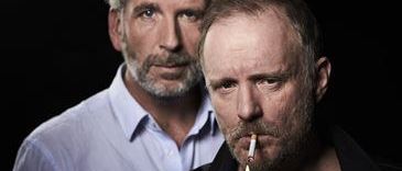 Zwei Männer stehen nebeneinander. Der Mann auf der linken Seite hat einen Bart und trägt ein hellblaues Hemd. Der Mann auf der rechten Seite raucht eine Zigarette, trägt ein schwarzes Hemd und hat einen strengen Ausdruck.