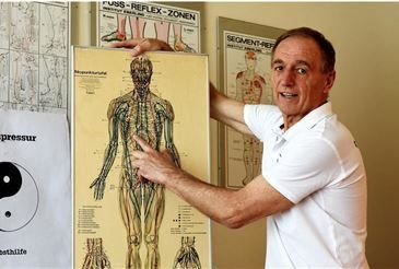 Ein Mann in einem weißen Poloshirt hält ein Diagramm des menschlichen Körpers hoch und steht neben Postern mit Akupunkturkarten und einem Yin-Yang-Symbol.
