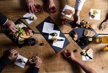 Mehrere Personen versammeln sich um einen Holztisch, der mit Essen und Getränken gefüllt ist. Der Tisch hat Käse, Trauben und andere Lebensmittel auf schwarzen Tellern. Die Leute halten Gläser mit Getränken und Besteck. Eine Flasche Alkohol steht ebenfalls auf dem Tisch.