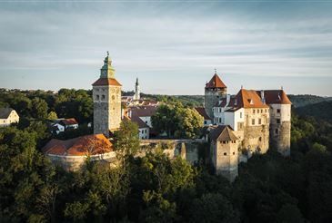 Ein Luftbild einer historischen Burg, umgeben von üppigem Grün, mit zwei markanten Türmen und einem roten Dach, vor einem bewölkten Himmel.