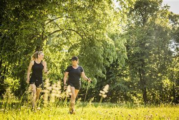 Zwei Frauen in Sportbekleidung wandern in einem Grasfeld mit Bäumen. Eine trägt ein schwarzes Shirt und Shorts, die andere ein ärmelloses Shirt. Beide benutzen Trekkingstöcke.