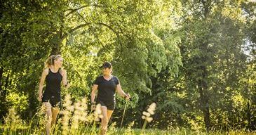 Zwei Frauen in Sportbekleidung wandern in einem Grasfeld mit Bäumen. Eine trägt ein schwarzes Shirt und Shorts, die andere ein ärmelloses Shirt. Beide benutzen Trekkingstöcke.