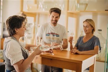 Drei Personen versammeln sich um einen Holztisch in einem gut beleuchteten Raum. Die Frau auf der linken Seite hält eine Flasche und ein Glas, während der Mann in der Mitte und die Frau auf der rechten Seite Gläser halten. Hinter ihnen sind mit Glaswaren gefüllte Regale zu sehen.