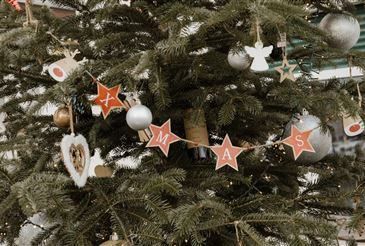 Ein Weihnachtsbaum ist mit Ornamenten geschmückt, darunter Sterne, Herzen und Engelsfiguren. Ein roter Banner mit weißen Buchstaben buchstabiert 'MAMA'. Der Baum steht in einem Raum mit einer grünen Wand.