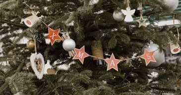 Ein geschmückter Weihnachtsbaum mit einer Banner, der 'M A S' lautet. Der Baum ist mit Ornamenten, Sternen und Engeln geschmückt. Es gibt auch herzförmige Dekorationen und einen kleinen Engel. Der Baum steht vor einem weißen Hintergrund.